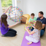 Karen showing the breathing sphere to a man and two children as they do the copy her sat on a yoga mat.