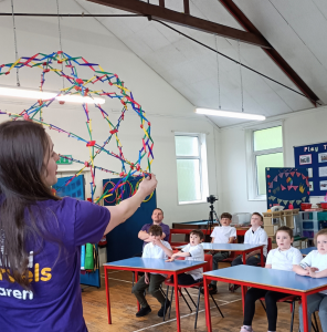 A woman with a purple t-shirt that says Mind Marvels holding up a contractable sphere in front of a classroom of children all watching her.