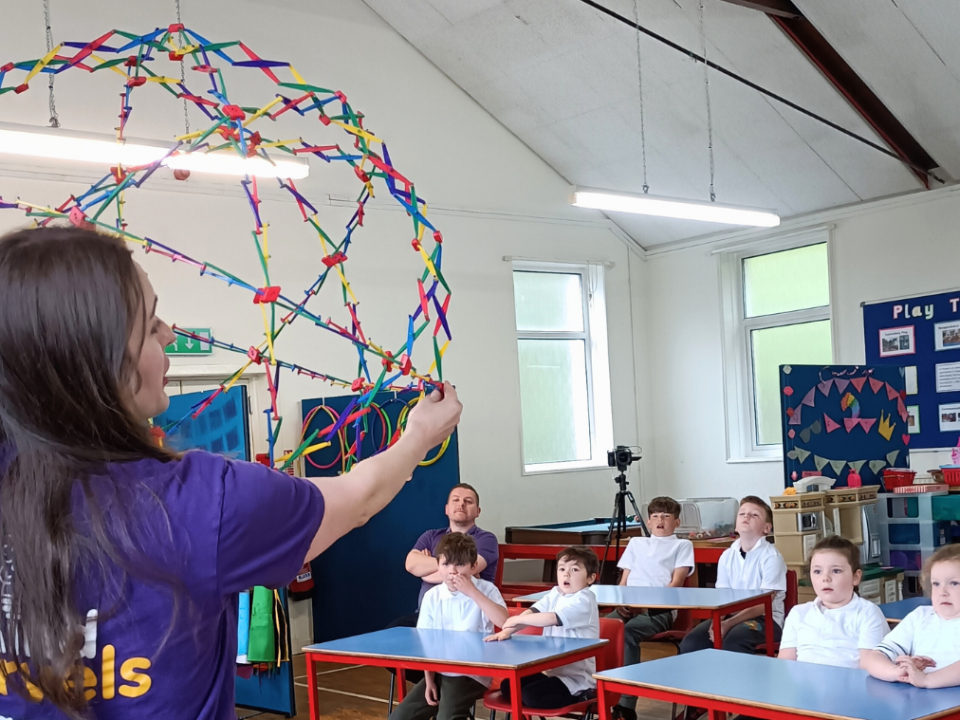 A woman with a purple t-shirt that says Mind Marvels holding up a contractable sphere in front of a classroom of children all watching her.