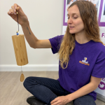 "A Mind Marvels instructor wearing a purple branded T-shirt sits cross-legged on the floor, gently holding and demonstrating a bamboo chime instrument during a wellbeing session."