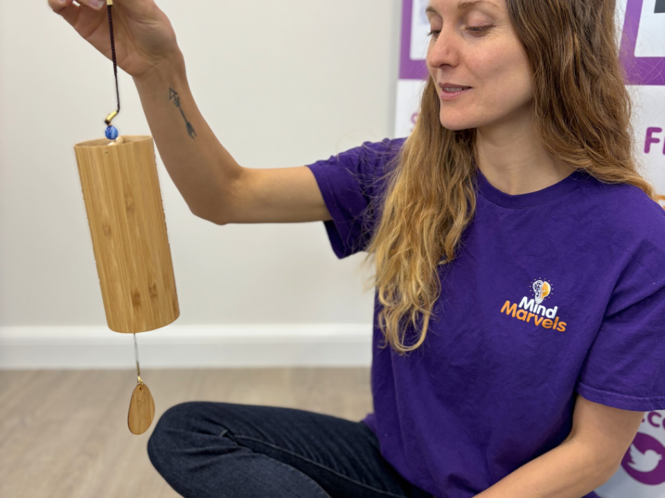"A Mind Marvels instructor wearing a purple branded T-shirt sits cross-legged on the floor, gently holding and demonstrating a bamboo chime instrument during a wellbeing session."